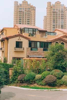 A residential area featuring a house with a beige facade and reddish-brown roof tiles, surrounded by lush greenery and shrubs. In the background, there are tall, modern apartment buildings with numerous windows and balconies, contributing to an urban yet peaceful setting.