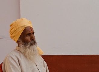 A man with a long white beard is sitting against a plain wall. He is wearing a pale orange turban and a light-colored shirt, perhaps indicative of traditional attire. The wall in the background is divided into a white upper section and a brown lower section.