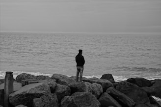 Wide shot of a solitary figure standing on a rocky shore beneath a cloudy sky, captured in black and white.