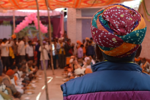 A person wearing a colorful, patterned turban and a blue jacket is facing a crowd. The crowd consists of people gathered, possibly at a cultural or festive event, with decorations like pink balloons and an orange canopy overhead. Many individuals appear to be seated on the floor, engaging in some communal activity.