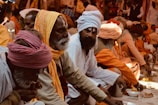A group of people in traditional attire are sitting closely together. They are dressed in orange, white, and beige robes with various head coverings like turbans and shawls. The setting appears to be a communal gathering, possibly involving religious or cultural practices. Some individuals are engaged in eating or preparing food placed in front of them.