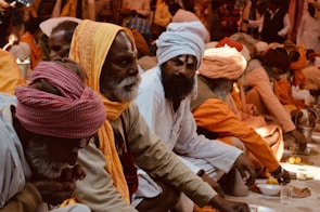 A group of people in traditional attire are sitting closely together. They are dressed in orange, white, and beige robes with various head coverings like turbans and shawls. The setting appears to be a communal gathering, possibly involving religious or cultural practices. Some individuals are engaged in eating or preparing food placed in front of them.