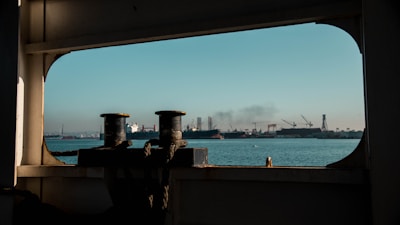 A view through a ship's window shows the sea with several large industrial ships and a bustling port in the distance. Cranes and other industrial structures are visible against a clear blue sky, and there's a hint of smoke rising from one of the ships, suggesting ongoing activity.