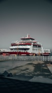 A large ferry is docked at a port, featuring multiple decks and a prominent red and white color scheme. The ship has a Turkish flag atop a mast, and the area is surrounded by metal fencing. The ground appears wet, suggesting recent rain.
