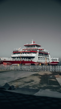 A large ferry is docked at a port, featuring multiple decks and a prominent red and white color scheme. The ship has a Turkish flag atop a mast, and the area is surrounded by metal fencing. The ground appears wet, suggesting recent rain.