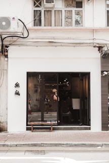 A street-level view of a building facade featuring a minimalist storefront with large glass doors and windows. The interior is visible with people inside, and there is a wooden bench placed on the sidewalk outside. The building has an air conditioning unit and wiring attached to the upper part above the storefront. Chinese characters are displayed on the white exterior wall to the left of the entrance.