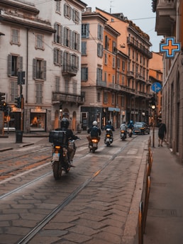 A group of motorcyclists rides along a narrow, cobblestone street lined with historic European-style buildings. The buildings feature ornate balconies and shuttered windows. The street is equipped with tram tracks, and a pharmacy cross sign glows on the right, indicating a pharmacy nearby. Pedestrians are visible on the sidewalk, and there’s a mix of old-world charm and modern city life.