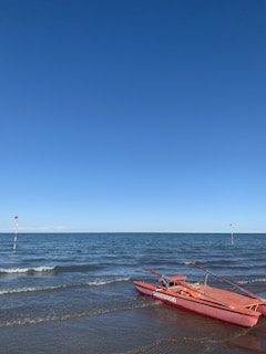 A red lifeguard boat labeled 'salvataggio' rests on a sandy beach near the water's edge. The ocean is calm with small waves, and the sky is clear with a vibrant blue hue. Two poles with red flags mark a boundary in the water.