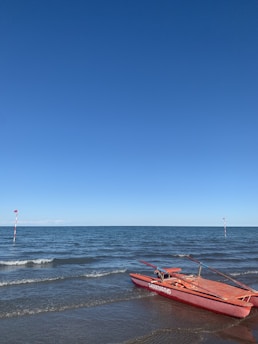 A red lifeguard boat labeled 'salvataggio' rests on a sandy beach near the water's edge. The ocean is calm with small waves, and the sky is clear with a vibrant blue hue. Two poles with red flags mark a boundary in the water.