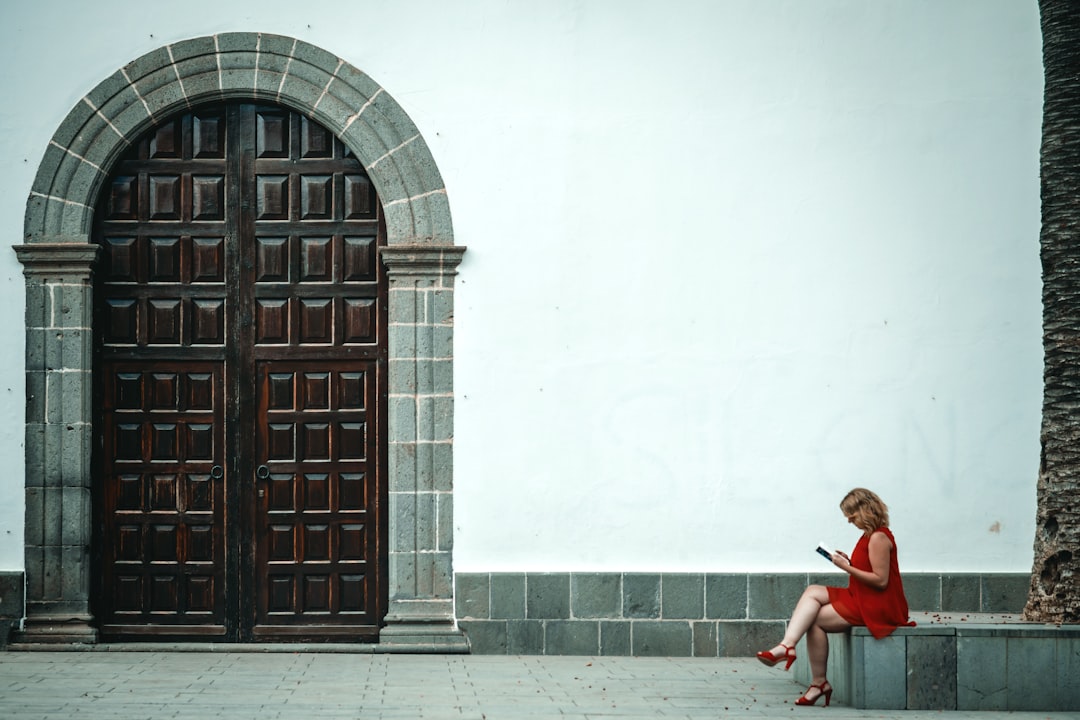 woman in red dress sitting on concrete bench near brown wooden door during daytime