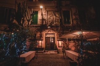 A rustic facade of an old building in a dimly lit street with a hanging streetlamp casting a warm glow. The entrance door labeled 'DA MEo PATacca' is flanked by green plants and small potted shrubs. Tables with checked tablecloths are positioned outside, suggesting a dining area. Twinkling lights are wrapped around nearby plants, adding a festive touch to the scene. An umbrella canopy slightly illuminated from beneath adds to the outdoor ambience.