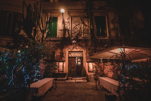 A rustic facade of an old building in a dimly lit street with a hanging streetlamp casting a warm glow. The entrance door labeled 'DA MEo PATacca' is flanked by green plants and small potted shrubs. Tables with checked tablecloths are positioned outside, suggesting a dining area. Twinkling lights are wrapped around nearby plants, adding a festive touch to the scene. An umbrella canopy slightly illuminated from beneath adds to the outdoor ambience.