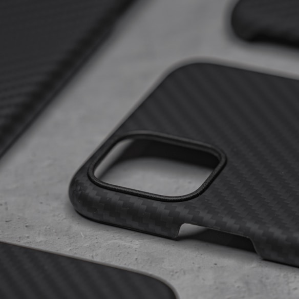 A close-up view of a carbon fiber textured phone case on a gray surface, featuring a cutout for the camera. The case has a sleek and minimalist design.