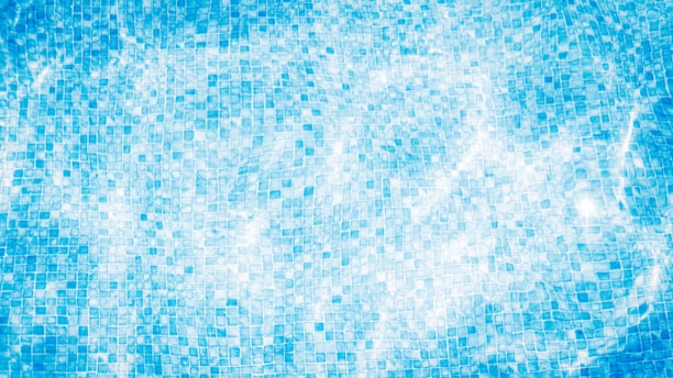 An overhead view of a swimming pool surface with a mosaic tile pattern visible through the water. The sunlight creates shimmering effects on the rippling water.