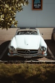 A classic white convertible car is parked in a parking space under a tree, with sunlight casting shadows on the hood. The car features an emblem on the front and a license plate. The background includes a sign for a supermarket.