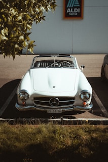 A classic white convertible car is parked in a parking space under a tree, with sunlight casting shadows on the hood. The car features an emblem on the front and a license plate. The background includes a sign for a supermarket.