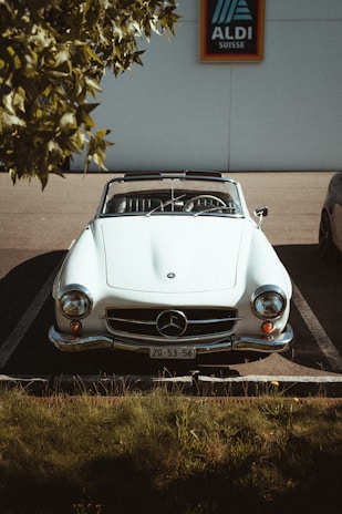 A classic white convertible car is parked in a parking space under a tree, with sunlight casting shadows on the hood. The car features an emblem on the front and a license plate. The background includes a sign for a supermarket.
