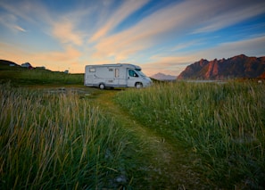 A camper van is parked on a grassy field with tall mountains in the background. The sky is a mix of blue and orange hues, suggesting a sunset or sunrise. In the distance, a couple of small houses or buildings are visible under the wide expanse of the sky.