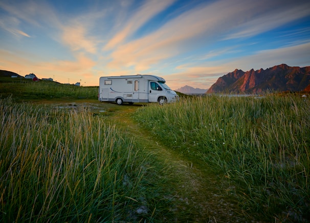 A camper van is parked on a grassy field with tall mountains in the background. The sky is a mix of blue and orange hues, suggesting a sunset or sunrise. In the distance, a couple of small houses or buildings are visible under the wide expanse of the sky.