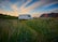 A camper van is parked on a grassy field with tall mountains in the background. The sky is a mix of blue and orange hues, suggesting a sunset or sunrise. In the distance, a couple of small houses or buildings are visible under the wide expanse of the sky.