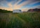 A camper van is parked on a grassy field with tall mountains in the background. The sky is a mix of blue and orange hues, suggesting a sunset or sunrise. In the distance, a couple of small houses or buildings are visible under the wide expanse of the sky.