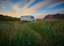 A camper van is parked on a grassy field with tall mountains in the background. The sky is a mix of blue and orange hues, suggesting a sunset or sunrise. In the distance, a couple of small houses or buildings are visible under the wide expanse of the sky.