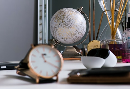 A desk is adorned with a small, gold-accented globe, an hourglass, and a variety of decorative and functional items, including a stylish watch in the foreground. A bottle of fragrance with thin reeds protruding, along with other bottles, adds a touch of elegance. The setting is neat, suggesting a study or personal workspace.