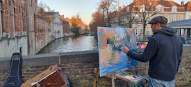 A man is painting an impressionist-style canvas by a canal lined with traditional European buildings under a clear sky. He wears casual outdoor clothing and a flat cap, and is focused on adding details to the vibrant artwork. The scene includes a scenic view of the waterway with reflections of the surrounding architecture.
