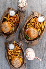 Three meals are arranged on a wooden table, each consisting of a burger with sesame seed buns, accompanied by a mix of regular and sweet potato fries. Each serving comes with a small cup of white dipping sauce. Two milkshakes, one topped with whipped cream and chocolate sprinkles, and the other with a pink straw, are placed between the meals.