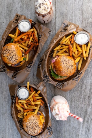 Three meals are arranged on a wooden table, each consisting of a burger with sesame seed buns, accompanied by a mix of regular and sweet potato fries. Each serving comes with a small cup of white dipping sauce. Two milkshakes, one topped with whipped cream and chocolate sprinkles, and the other with a pink straw, are placed between the meals.