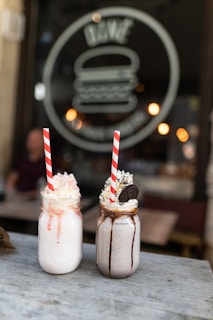Two mason jar milkshakes with whipped cream and striped paper straws sit on a wooden table. The left jar appears to have a strawberry flavor with drips along the side, while the right jar is chocolate with cookie pieces and a small chocolate cookie garnish. The background features a blurred view of a café setting with a large logo in the window and warm ambient lighting.
