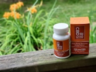 A white bottle and a box of CBD gel capsules are placed on a wooden surface. The bottle has a white cap and orange labeling, while the orange box is placed upright next to it. The background features blurry greenery and orange flowers, suggesting an outdoor setting.