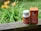 A white bottle and a box of CBD gel capsules are placed on a wooden surface. The bottle has a white cap and orange labeling, while the orange box is placed upright next to it. The background features blurry greenery and orange flowers, suggesting an outdoor setting.