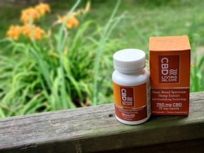A white bottle and a box of CBD gel capsules are placed on a wooden surface. The bottle has a white cap and orange labeling, while the orange box is placed upright next to it. The background features blurry greenery and orange flowers, suggesting an outdoor setting.