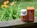 A white bottle and a box of CBD gel capsules are placed on a wooden surface. The bottle has a white cap and orange labeling, while the orange box is placed upright next to it. The background features blurry greenery and orange flowers, suggesting an outdoor setting.