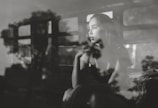 A contemplative woman gazes out of a window, with reflections of trees and a window frame creating an artistic overlay. The scene is captured in black and white, enhancing the depth of her thoughtful expression.
