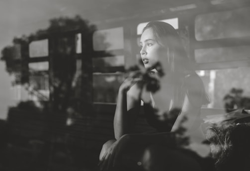 A contemplative woman gazes out of a window, with reflections of trees and a window frame creating an artistic overlay. The scene is captured in black and white, enhancing the depth of her thoughtful expression.