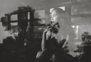 A contemplative woman gazes out of a window, with reflections of trees and a window frame creating an artistic overlay. The scene is captured in black and white, enhancing the depth of her thoughtful expression.