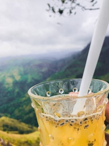 A glass filled with a yellow-orange beverage containing chia or basil seeds, held near a scenic view of lush green hills and a cloudy sky. A straw is placed in the glass.