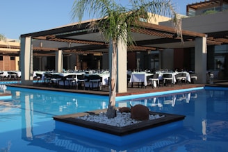 An outdoor dining area with several tables neatly arranged under a wooden pergola, surrounded by a clear, blue swimming pool. Each table is covered with white tablecloths and set for a meal. A small, landscaped island in the pool features a palm tree and some decorative rocks.