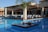 An outdoor dining area with several tables neatly arranged under a wooden pergola, surrounded by a clear, blue swimming pool. Each table is covered with white tablecloths and set for a meal. A small, landscaped island in the pool features a palm tree and some decorative rocks.
