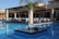 An outdoor dining area with several tables neatly arranged under a wooden pergola, surrounded by a clear, blue swimming pool. Each table is covered with white tablecloths and set for a meal. A small, landscaped island in the pool features a palm tree and some decorative rocks.