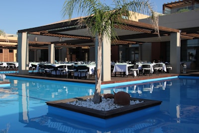 An outdoor dining area with several tables neatly arranged under a wooden pergola, surrounded by a clear, blue swimming pool. Each table is covered with white tablecloths and set for a meal. A small, landscaped island in the pool features a palm tree and some decorative rocks.