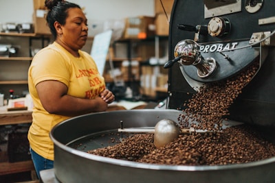 A person wearing a yellow shirt stands next to a coffee roasting machine, which is releasing roasted coffee beans into a large circular tray. The setting appears to be a coffee roasting facility with various equipment and shelves in the background.