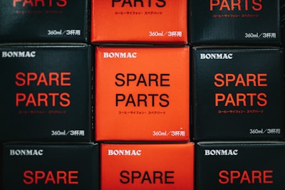 A collection of boxed items stacked neatly, featuring contrasting colors of black and red. The text 'SPARE PARTS' is prominently displayed on each box, accompanied by smaller text and brand name 'BONMAC'.