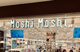 A retail store with bright lighting and organized displays featuring a variety of products. The signage 'Moshi Moshi' is prominently displayed above the entrance. The store interior includes sections with shelves filled with colorful merchandise such as gadgets, accessories, and home goods. TV screens are mounted on the wall, displaying product advertisements.