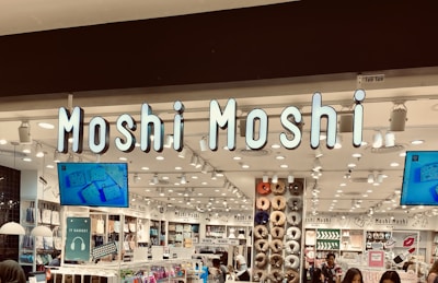 A retail store with bright lighting and organized displays featuring a variety of products. The signage 'Moshi Moshi' is prominently displayed above the entrance. The store interior includes sections with shelves filled with colorful merchandise such as gadgets, accessories, and home goods. TV screens are mounted on the wall, displaying product advertisements.