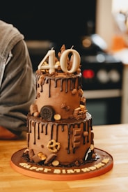 A two-tiered chocolate cake adorned with various chocolates and a '40' number candle on top. The cake features a dripped chocolate ganache design and is set on a wooden table with a slight view of a kitchen in the background.