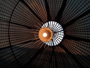 A close-up view of a light fixture with a grid-like pattern casting geometric shadows around a glowing central bulb. The design forms a circular pattern that radiates outward, creating a dynamic play of light and shadow.