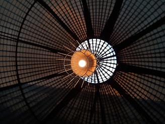 A close-up view of a light fixture with a grid-like pattern casting geometric shadows around a glowing central bulb. The design forms a circular pattern that radiates outward, creating a dynamic play of light and shadow.